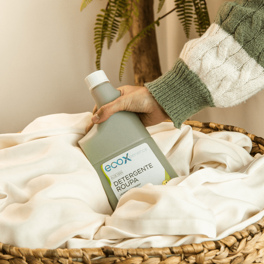Mão a agarrar numa embalagem de detergente roupa aloe vera EcoX 850mL, que está em cima de um cesto com roupa