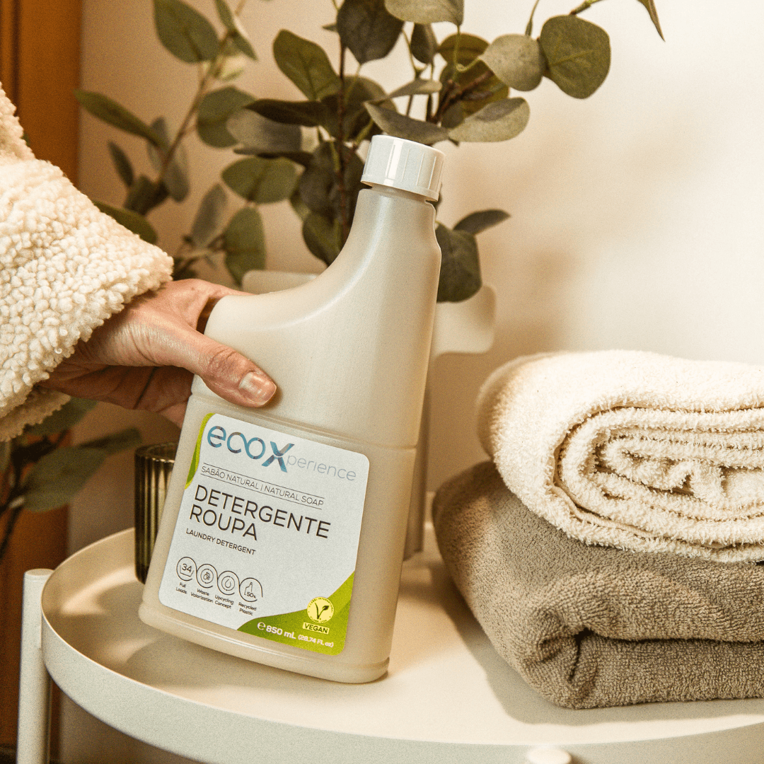 Mão a segurar uma embalagem de detergente roupa sabão natural EcoX 850mL que está ao lado de toalhas