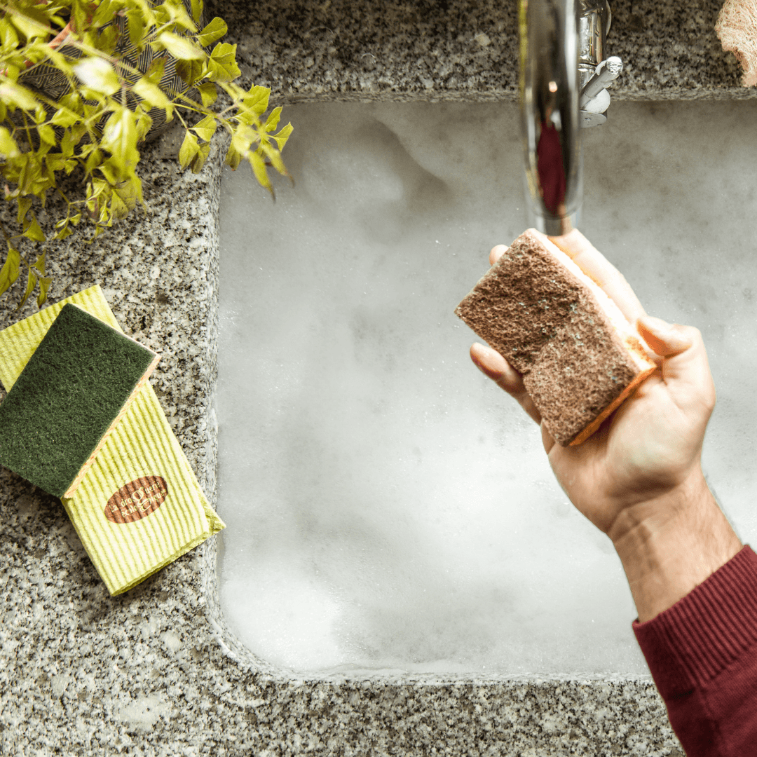 Mão a segurar na esponja da loiça com a esponja de celulose ao lado da pia em cima da bancada