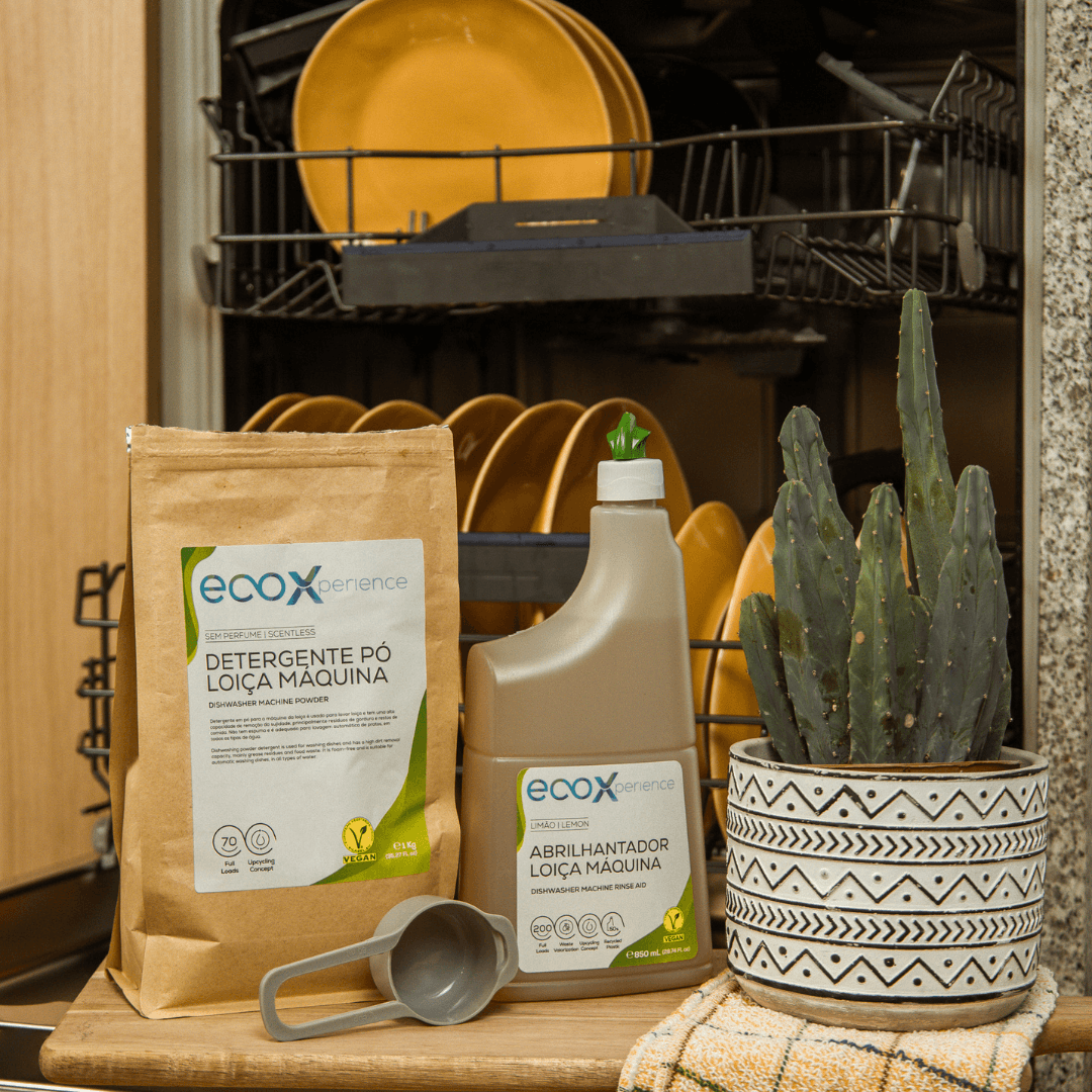 Abrilhantador para máquina de lavar loiça ao lado do detergente loiça máquina e de uma planta com a máquina de lavar loiça atrás