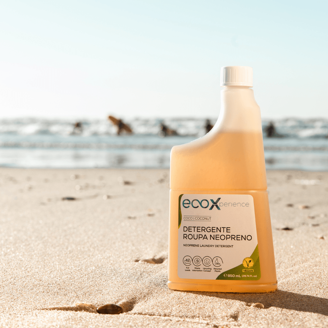 Embalagem de detergente neopreno EcoX 850mL na areia com os surfistas atrás no mar