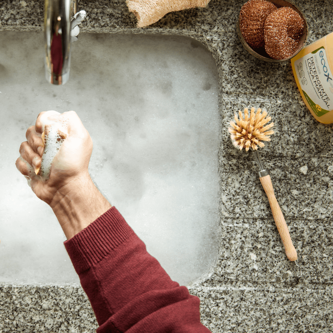 Mão a apertar a esponja da loiça com a recarga para escova de madeira na pega em cima da banca da cozinha